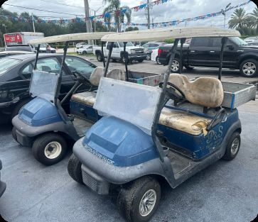 Two worn blue Club Car golf carts with damaged seats in a parking lot
